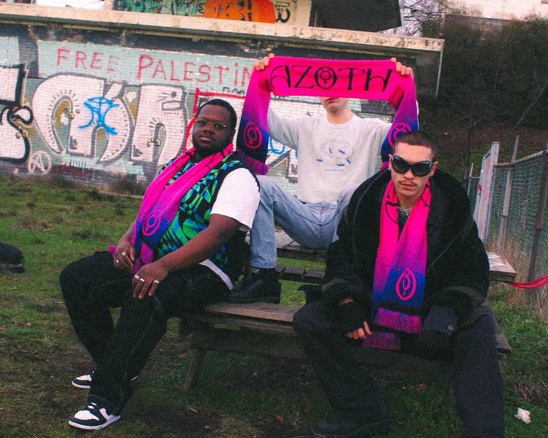 Three people on a bench holding up a pink and blue AZOTH scarf