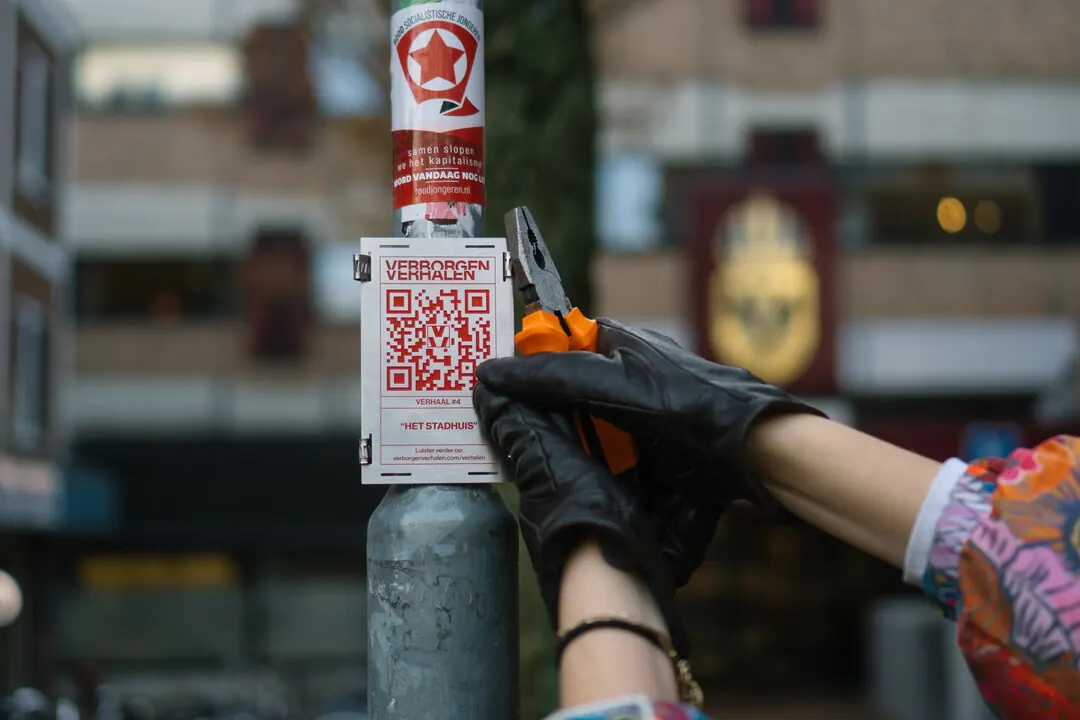 A gloved hand tying a small poster with a QR code to a lamppost