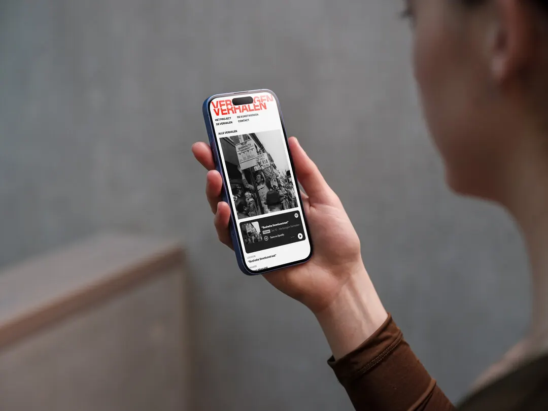 A hand holding a phone showing the Verborgen Verhalen website